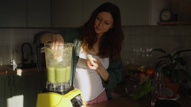 Expectant mother preparing a nutritious smoothie in her kitchen, adding fresh ingredients like bananas and vegetables into a blender for a healthy diet and proper nutrition during pregnancy