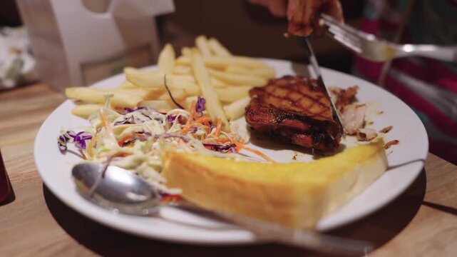 Grilled chicken steak served with crispy French fries, creamy coleslaw, and buttered toast on a white plate. Person using cutlery to cut meat at a restaurant. Savory and delicious food presentation.