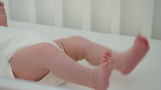 Baby lifts both legs and moves them energetically in crib, showing active wakefulness, natural body movement, and growing coordination during calm indoor care in early infancy at home.