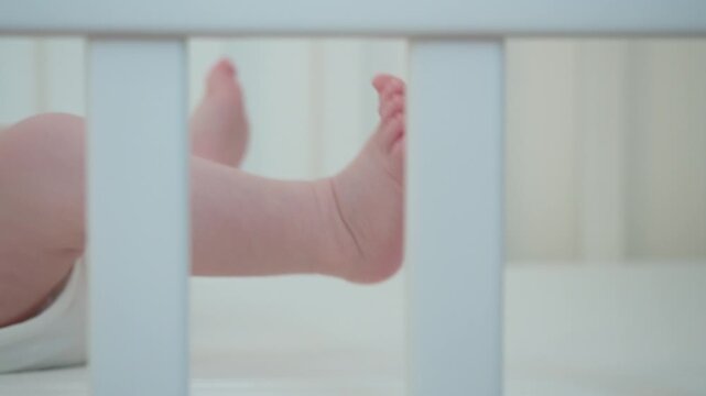 Baby moves legs behind crib bars, creating visual image of protected home space where everyday care, safe rest, and natural physical activity come together during early infancy indoors.
