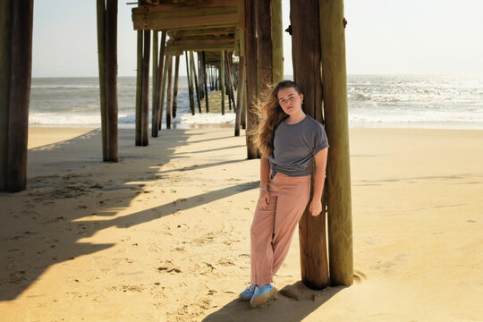 A young woman with long hair leans against a pier post on the beach