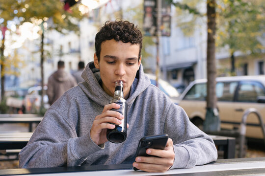 Young man drinks a soda while checking his phone at a street cafe