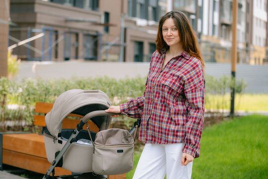 Young mother smiling, holding baby carriage handle while walking outdoors in modern city park, enjoying motherhood and a comfortable lifestyle