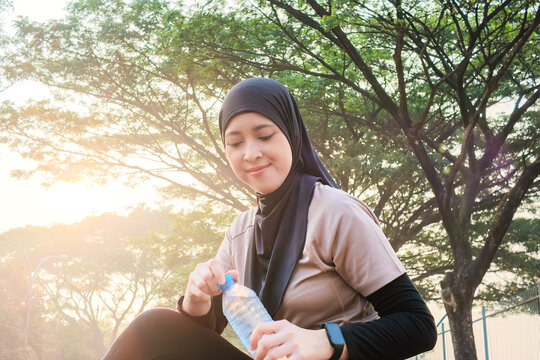 Smiling Asian Muslim woman pauses and taking a Water Break during an outdoor fitness session or jogging session in a sunlit park, hydrating with a water bottle and enjoying a calm, healthy moment afte