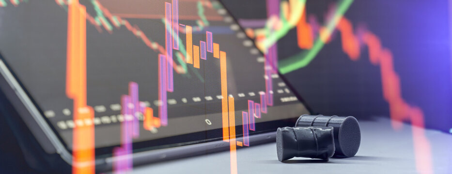 Crude oil price drop concept. Miniature black barrels lying on a table against glowing financial stock market charts