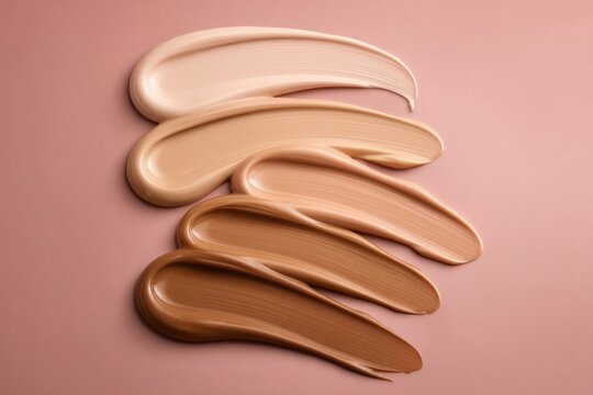 Collection of neutral and warm foundation makeup swatches in various shades, textures and brush strokes arranged in an artistic flat lay composition on a clean beige background.