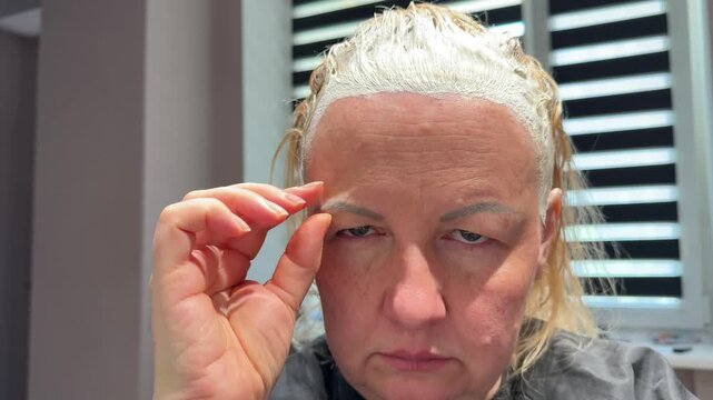 Mature woman undergoes hair bleaching treatment in salon while touching and lifting skin near her eye, reflecting concerns about aging, beauty and rejuvenation. Close up.