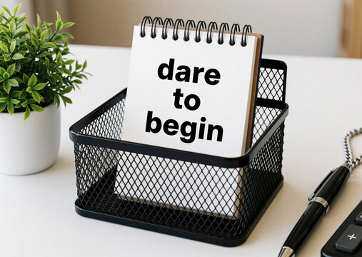 Notebook with dare to begin text sits on desk. Pen, plant, calculator nearby. Clean workspace offers inspiration for new goals and fresh starts.