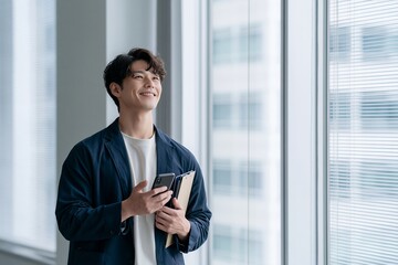 Fototapeta premium 笑顔で窓の外を眺めるスマートフォンを持つ若手ビジネスマン