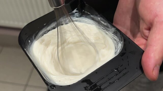 Male hairdresser hand mixes creamy hair coloring product with a whisk in a black bowl, preparing professional dye mixture before salon coloring procedure. Close up.