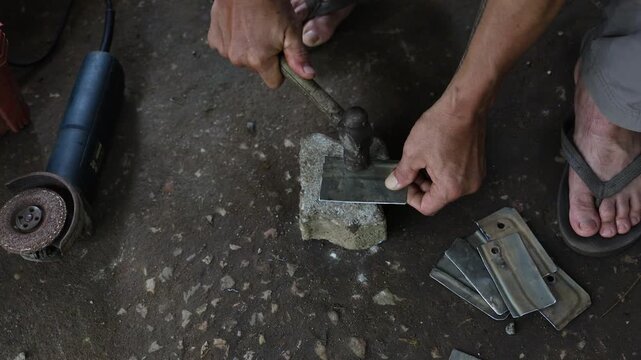 Metalworking artisan hammering hot metal on stone block with hand tools and grinding equipment, shaping rough surfaces through traditional workshop craftsmanship and manual forging labor