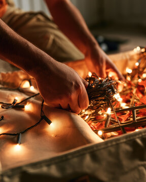 Adult hands coiling string light on canvas bag during seasonal decoration storage in warm indoor light, with tangled wire and glowing bulb creating careful domestic maintenance scene