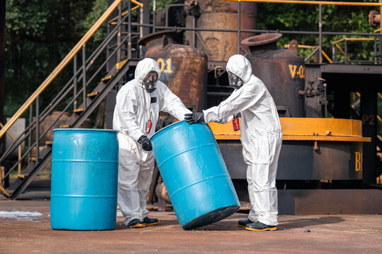 Hazmat workers tilting and moving a chemical storage drum together during hazardous materials response and workplace safety training at an outdoor industrial facility in protective suits