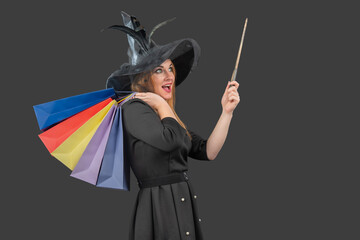 Young woman dressed as a witch with colorful shopping bags and a wand, casting a playful magic spell while pointing, excited about Halloween sales and festive shopping © somemeans
