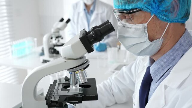 Senior male scientist wearing a protective mask, cap, and gloves, examining samples under a microscope in a modern laboratory. Medicine, healthcare and science concept