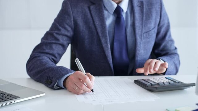 Businessman wearing suit and tie is using calculator and taking notes while working at desk in office, with laptop and stack of folders nearby, close up. Audit and taxes theme in business