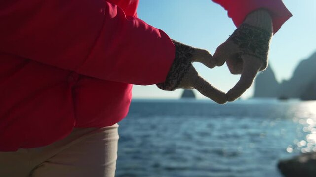 Heart gloves ocean woman wearing red jacket forms heart shape with hands by the sparkling sea on a sunny rocky coast