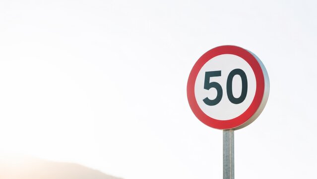 Round red-bordered speed limit fifty road sign mounted on a slim metal pole standing alone against a soft overcast sky with faint mountain silhouettes visible on the pale minimalist horizon