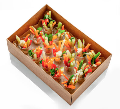 Fresh vegetable sticks served in individual cups. Sliced raw vegetables stand vertically in small transparent cups inside a takeaway box over a white surface.