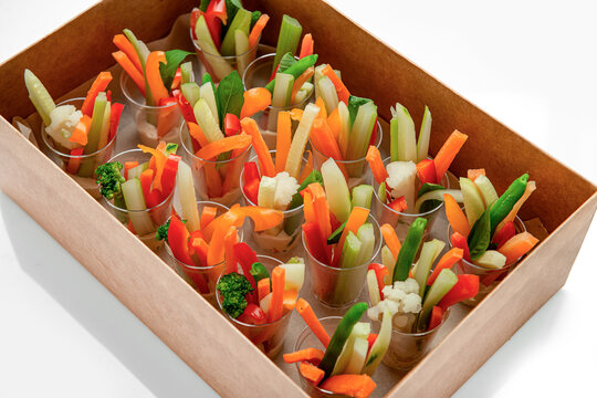 Fresh vegetable sticks in cups placed inside cardboard catering box. Sliced carrots, cucumbers, celery, and peppers stand in small transparent cups arranged in brown paper box.