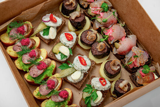 Assorted gourmet canapes arranged inside cardboard catering box. Small open sandwiches with sausage, fish, lard, and herbs are neatly placed in brown paper container.