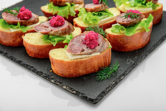 Sausage and beetroot spread canapes served on slate platter. Sliced baguette topped with cheese, sausage, red horseradish, and dill is arranged on black slate board.