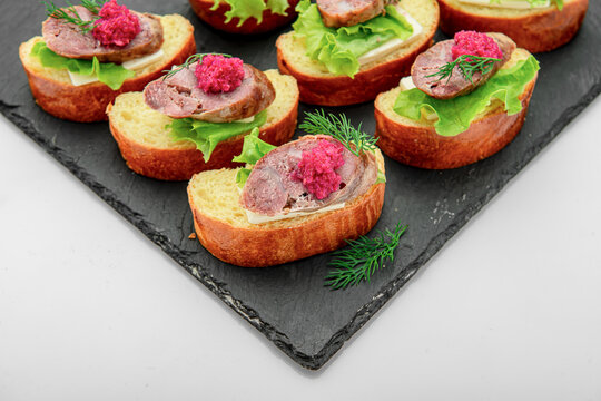 Crostini served with meat and salad. Small pieces of bread topped with meat, greens, and pink spread on a black platter. Ideal for a gathering or party.