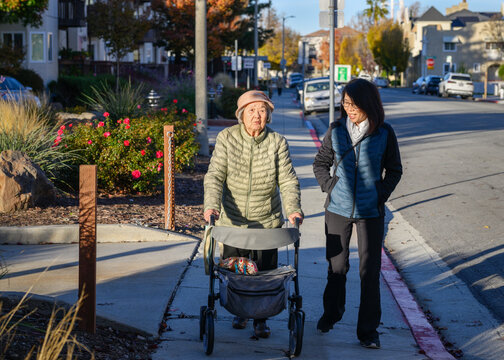 Senior woman, accompanied by adult daughter, walking using a mobility walker. Autumn sun casting long shadows. California.