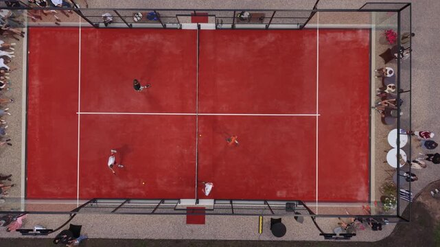 Four players engage in a competitive match on a red paddle tennis court surrounded by spectators enjoying the game
