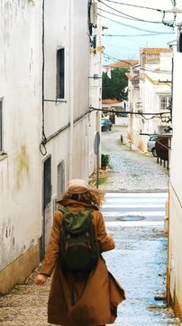 Back view of a young woman wearing a backpack and walking down a narrow, ancient cobblestone street in a traditional portuguese village, enjoying her vacation in europe