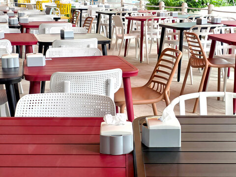 Rows of empty outdoor restaurant tables and chairs in red, black and white. Plastic furniture with napkin dispensers on a paved terrace. Modern casual dining area waiting for customers.