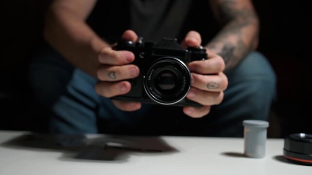 Tattooed man's hands carefully holding and examining a classic analog camera, preparing it for use with a film roll on a white table