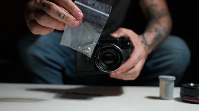 Tattooed male technician assembling a classic analog camera on a white desk. His hands carefully handle small parts from a plastic bag