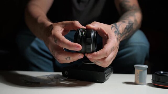 Close up of a tattooed person's hands carefully assembling a vintage analog camera lens. Film camera and roll visible in the background