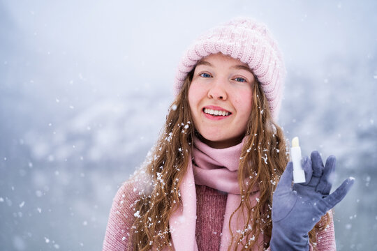 Friendly young woman in warm pink clothing smiling during a winter snowfall, looking at the camera and holding a lip balm. Beauty, skin care, winter lifestyle, and cold weather concepts.