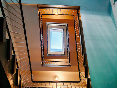 Stairwell geometry viewed upward toward a bright skylight, with repeating railings, warm interior lighting, and contrasting turquoise and orange walls creating a strong architectural composition