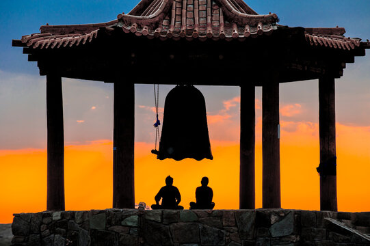 Die Silhouetten zweier meditierender Menschen unter einer Glocke eines buddhitischen Tempels bei stimmungsvollem, orangefarbenem Himmel, W&uuml;ste Gobi, Mongolei, Zentralasien