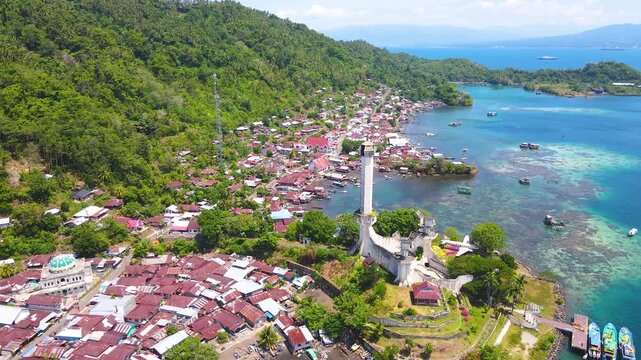 Aerial view of Batu Lubang, Lembeh Island in Bitung, Indonesia. The iconic Trikora Monument stands tall above a vibrant fishing village and boats moored in crystal-clear turquoise waters.