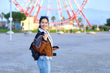 Naklejka premium Smiling traveler with backpack and camera at an outdoor park, ferris wheel in background, casual layered clothing, sunny day, adventure and exploration