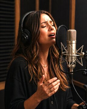 Woman with headphones sings into a studio microphone behind a pop filter, conveying focused concentration, creative expression, and audio recording.