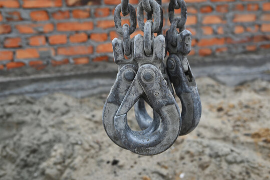 Close-up of heavy metal lifting hooks and chain links used for industrial hoisting and rigging, with a blurred brick wall and construction site in the background.