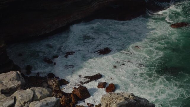 Turquoise ocean waves surge and break over dark coastal rocks, creating frothy white patterns and vivid green water in a rugged shoreline scene, with natural motion and textured seascape detail.