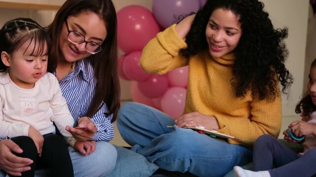 Two female teachers and little girls playing a learning game sitting on the floor in kindergarten