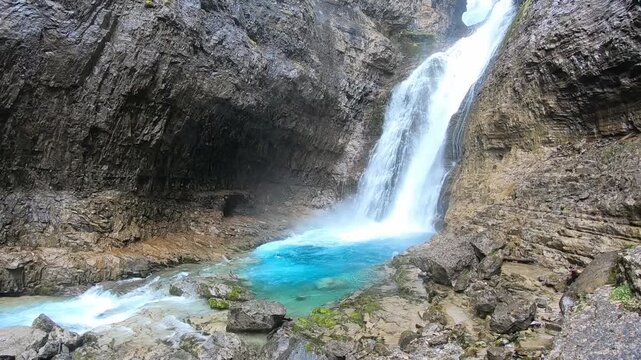 A tall waterfall cascades down a narrow rock canyon into a vivid turquoise pool. Steep layered rock walls frame the falling water, creating a dramatic natural scene carved by centuries of erosion.
