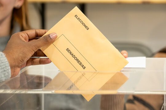 Hand inserting election envelope for senators into a transparent ballot box during voting