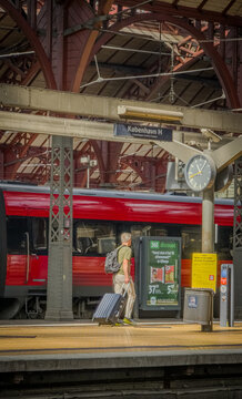 Copenhage, Denmark. August 15, 2026, Man with backpack and rolling luggage walking along the platform, a red train arriving at Copenhagen Central Station