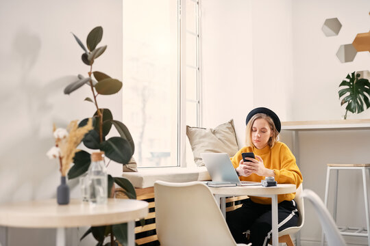 Woman using smart phone while working on laptop at cafe