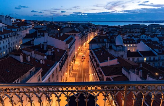Elevated dusk cityscape view of sao jorge street with glowing lights, ornate railing foreground, architectural buildings and horizon water at twilight generate ai
