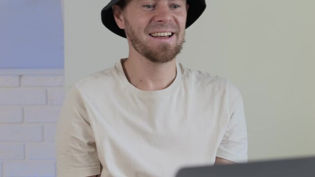 Young bearded man in black hat using game controller beside laptop, leaning forward with focused smile in casual indoor setting, showing relaxed posture and engaged expression