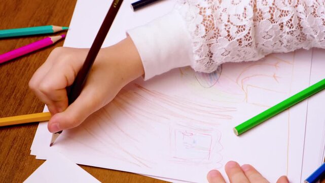 Young child hand sketches with pencil on lined paper, adding light color and shape. Bright pencils on wood table frame quiet drawing practice for school readiness and creativity through tracing lines.